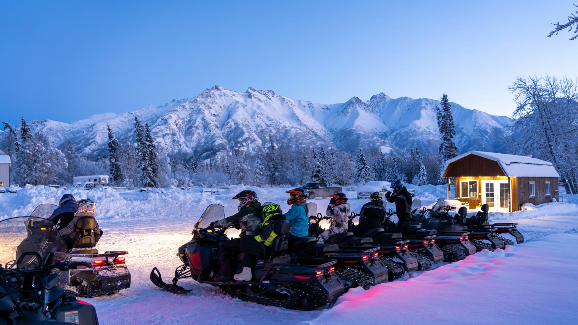 Snowmobile group lined up at dusk below snowy Chugach peaks